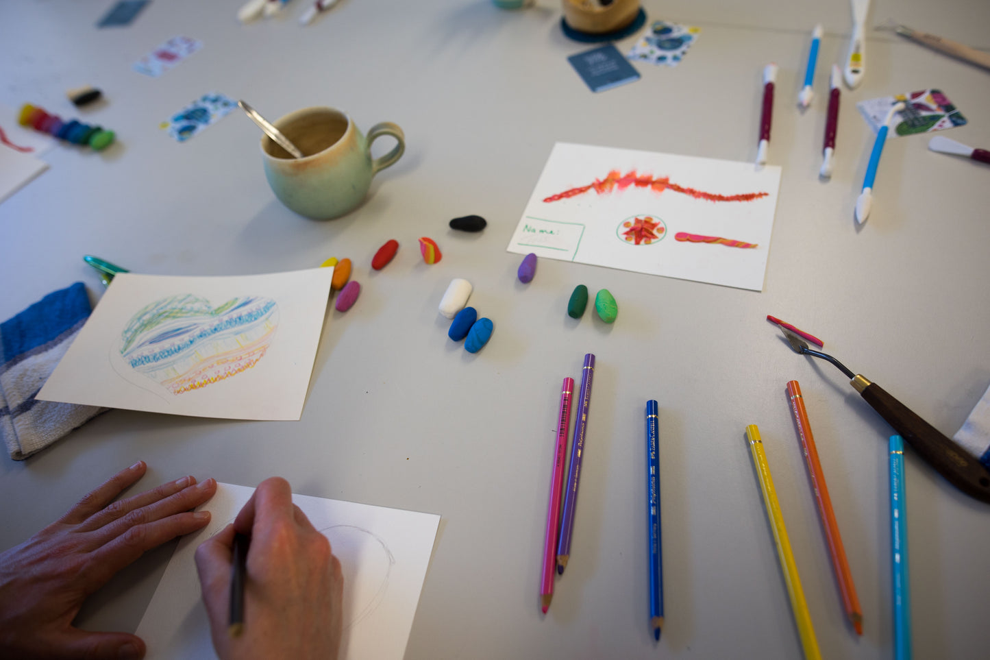 Beautiful hands of a Mindful Clay Art Workshop participant sketching her first non-dry clay art piece on a heart-shaped base, surrounded by a warm cacao drink, colorful pencils, clay, and modeling tools, embracing creativity and mindfulness.