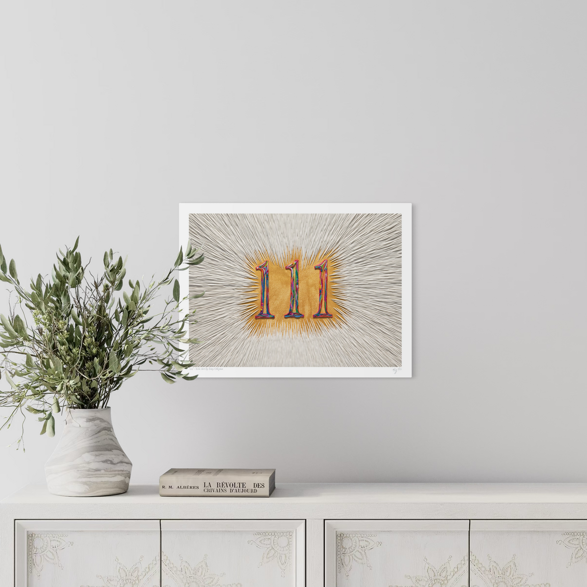 An A3 art print featuring the number 111, as an angel number, in a colorful, stylized plasticine piece against a golden acrylic paint background with radiating white lines, displayed on a light gray wall above a white cabinet with a vase of green leaves and a book. Art by Emy Oikawa from Eclo Art