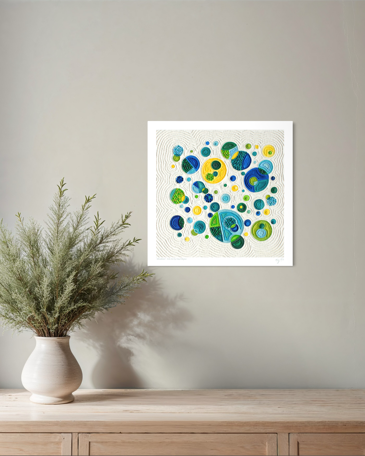 A square abstract wall art print called ''Abundance'' with colorful bubble-like circles in blue, green, yellow, and teal on a white background, displayed on a light gray wall above a wooden console table with a vase of greenery. Art by clay artist Emy Oikawa from Eclo Art.