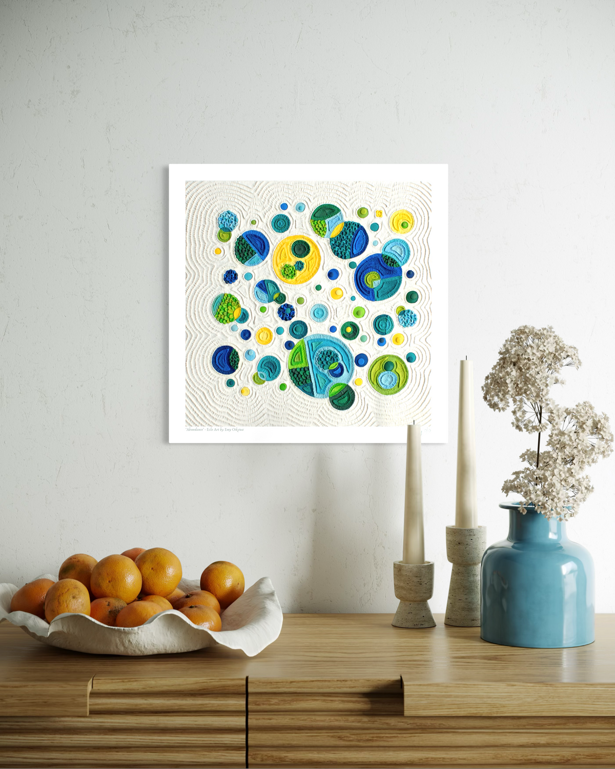 A square fine wall art print called ''Abundance'' featuring colorful abstract circles in blue, green, yellow on a cream plasticine colour background, displayed above a wooden console table with oranges, candles, and a blue vase in a styled interior setting. By New Zealand clay artist Emy Oikawa from Eclo Art