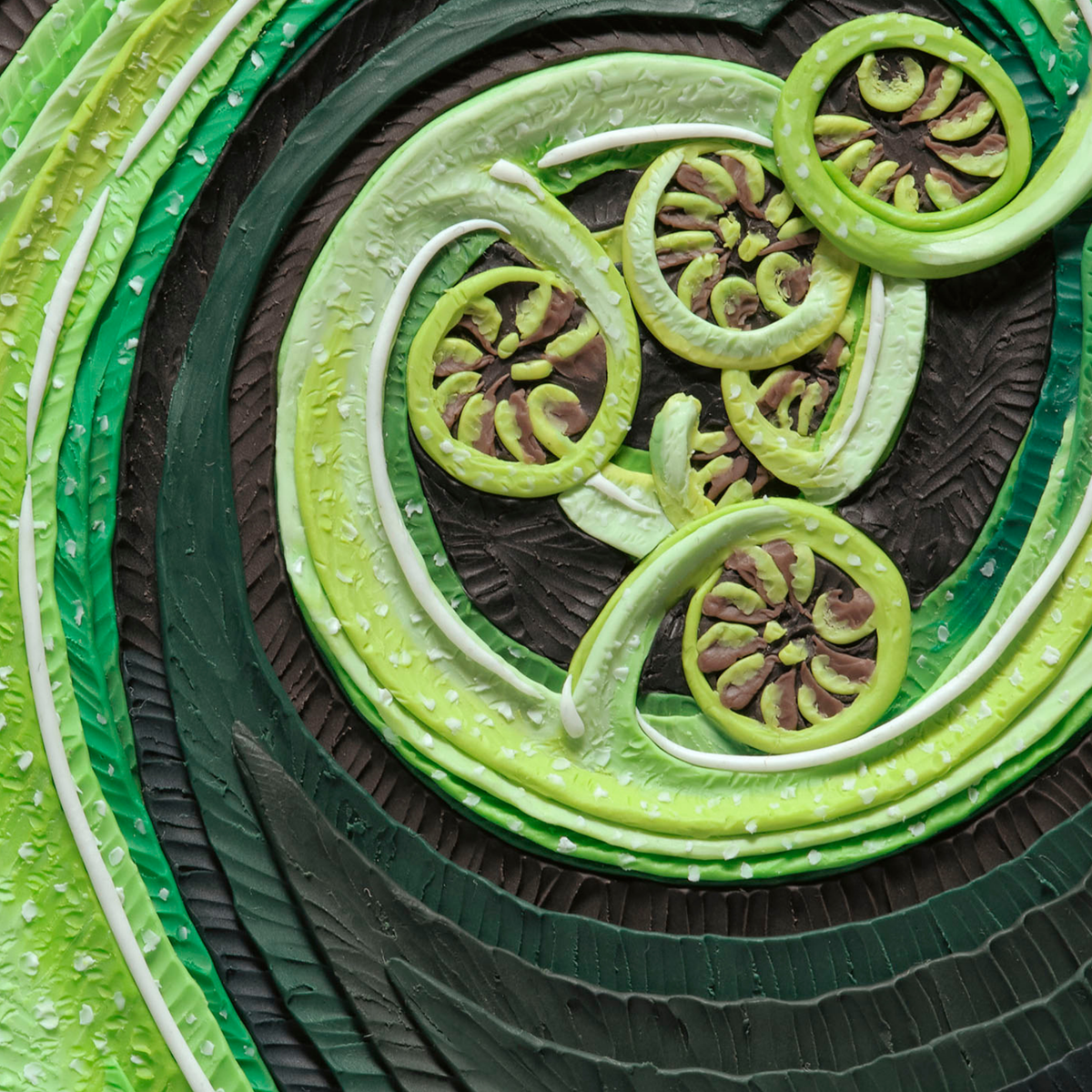 A close-up overhead view of a sculpted decorative art print called Mamaku, a New Zealand fern, wall art with layered green bands and circular green-and-brown or leaf-like motifs on a dark textured background. Art by clay artist Emy Oikawa from Eclo Art. New Zealand nature.