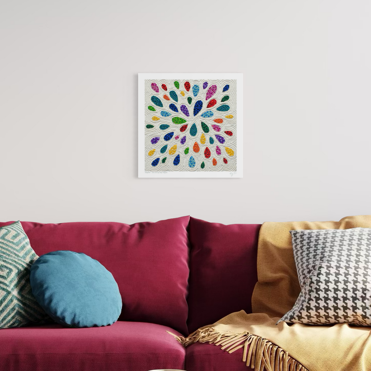 A square plasticine art print called ''Empower'' featuring a colorful radial burst of teardrop-like shapes on a light background, displayed on a white wall above a burgundy sofa with decorative pillows and a mustard throw blanket. Art by New Zealand Artist Emy Oikawa from Eclo Art.