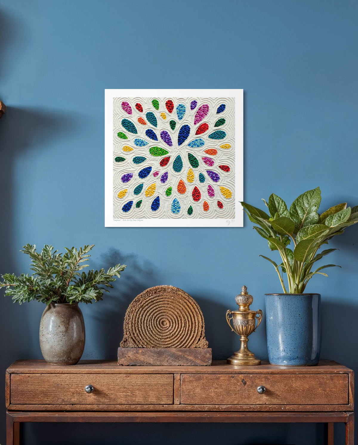 A square plasticine wall art print called ''Empower'' featuring a colorful radial pattern teardrop shapes on a white background, displayed on a blue wall above a wooden console table with plants and decorative objects. Art by New Zealand Artist Emy Oikawa from Eclo Art.