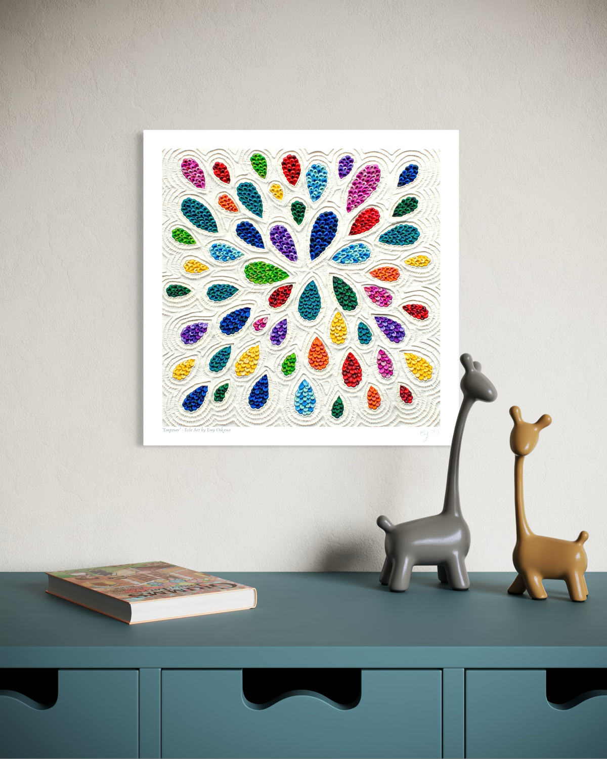 A square plasticine wall art print called ''Empower'' featuring a colorful radial pattern of teardrop shapes on a white background, displayed above a teal cabinet with decorative giraffe figurines and a book. Art by New Zealand Artist Emy Oikawa from Eclo Art.