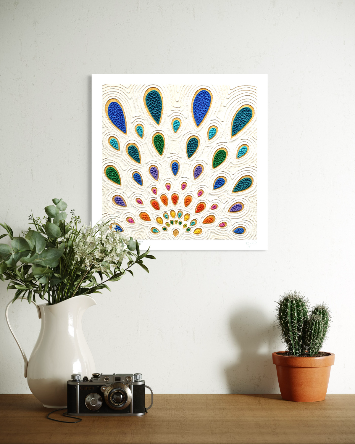 A square wall art print featuring a colorful peacock feather pattern-like called ''Growth'', hangs on a white wall above a wooden table styled with a white pitcher of greenery, a vintage camera, and a small potted cactus. Plasticine piece made by artist Emy Oikawa from Eclo Art.
