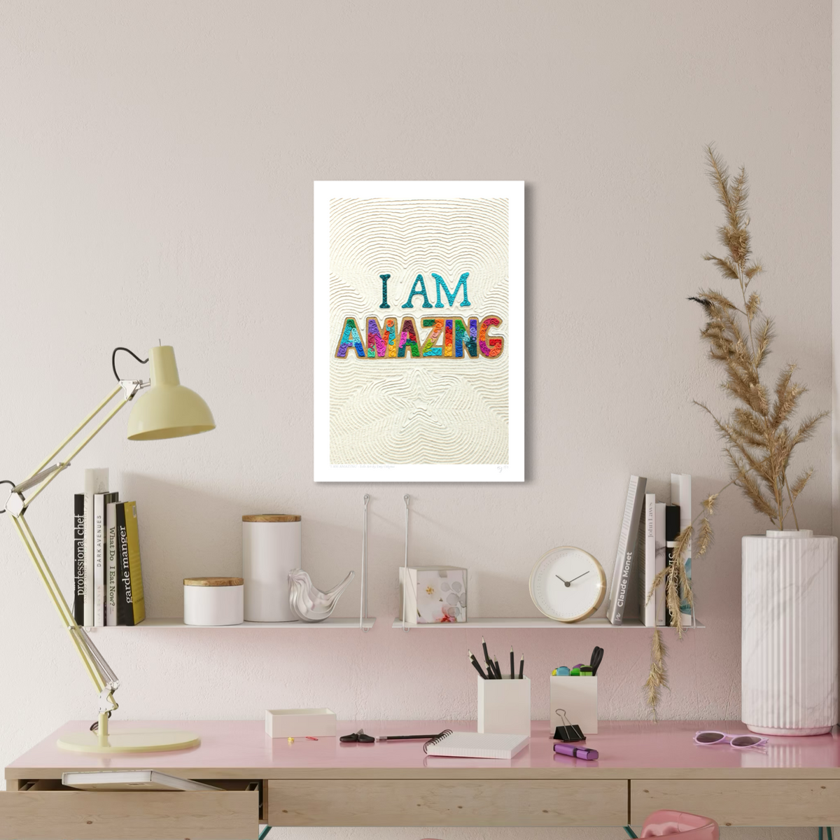 An A3 wall art print with the text 'I AM AMAZING' in colorful letters on a textured cream background with a start shape at the bottom, displayed on a beige wall above a pink desk with books, a lamp, and decorative items. Plasticine clay art by New Zealand artist Emy Oikawa from Eclo Art.