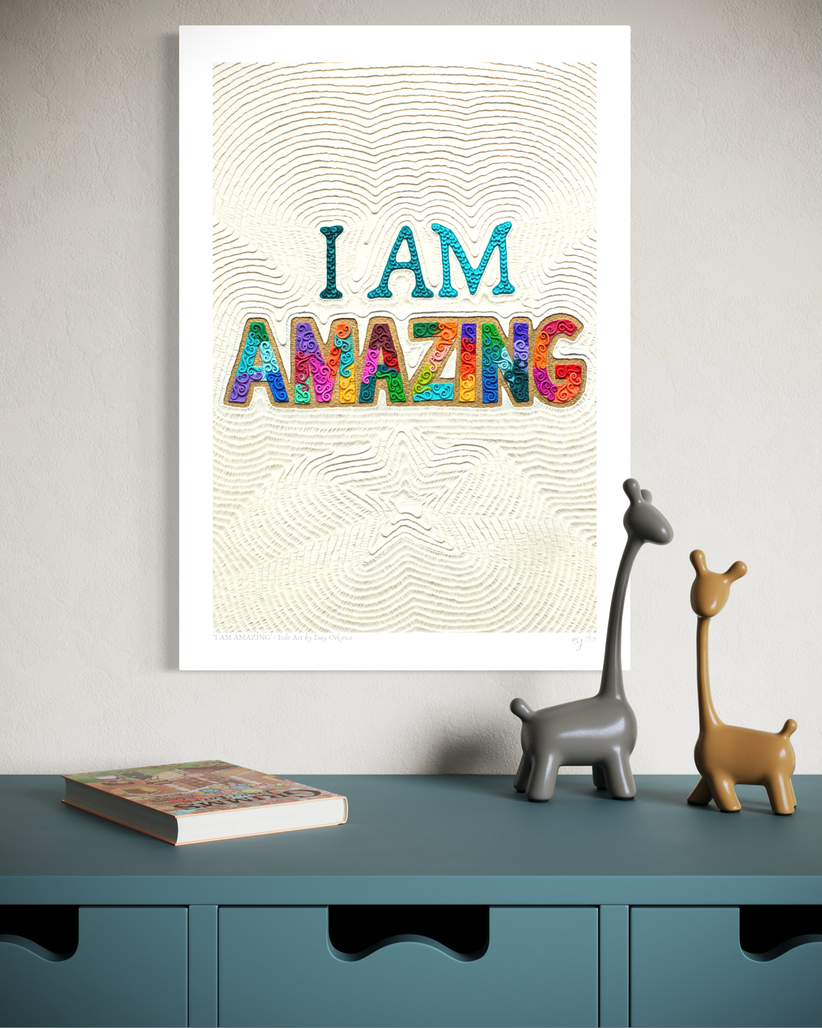 AN A3 wall art print with the words “I AM AMAZING” in colorful lettering hangs on a light gray wall above a teal cabinet. The print has a cream textured background with wavy line patterns. Plasticine clay art by New Zealand artist Emy Oikawa from Eclo Art.