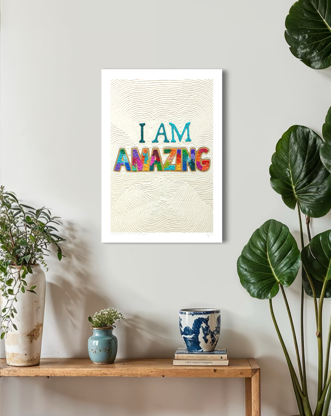 An A3 wall art print with the text 'I AM AMAZING' in colorful letters on a cream textured background with a star, displayed on a light gray wall above a wooden shelf with plants and small decor items. Plasticine clay art by New Zealand artist Emy Oikawa from Eclo Art.