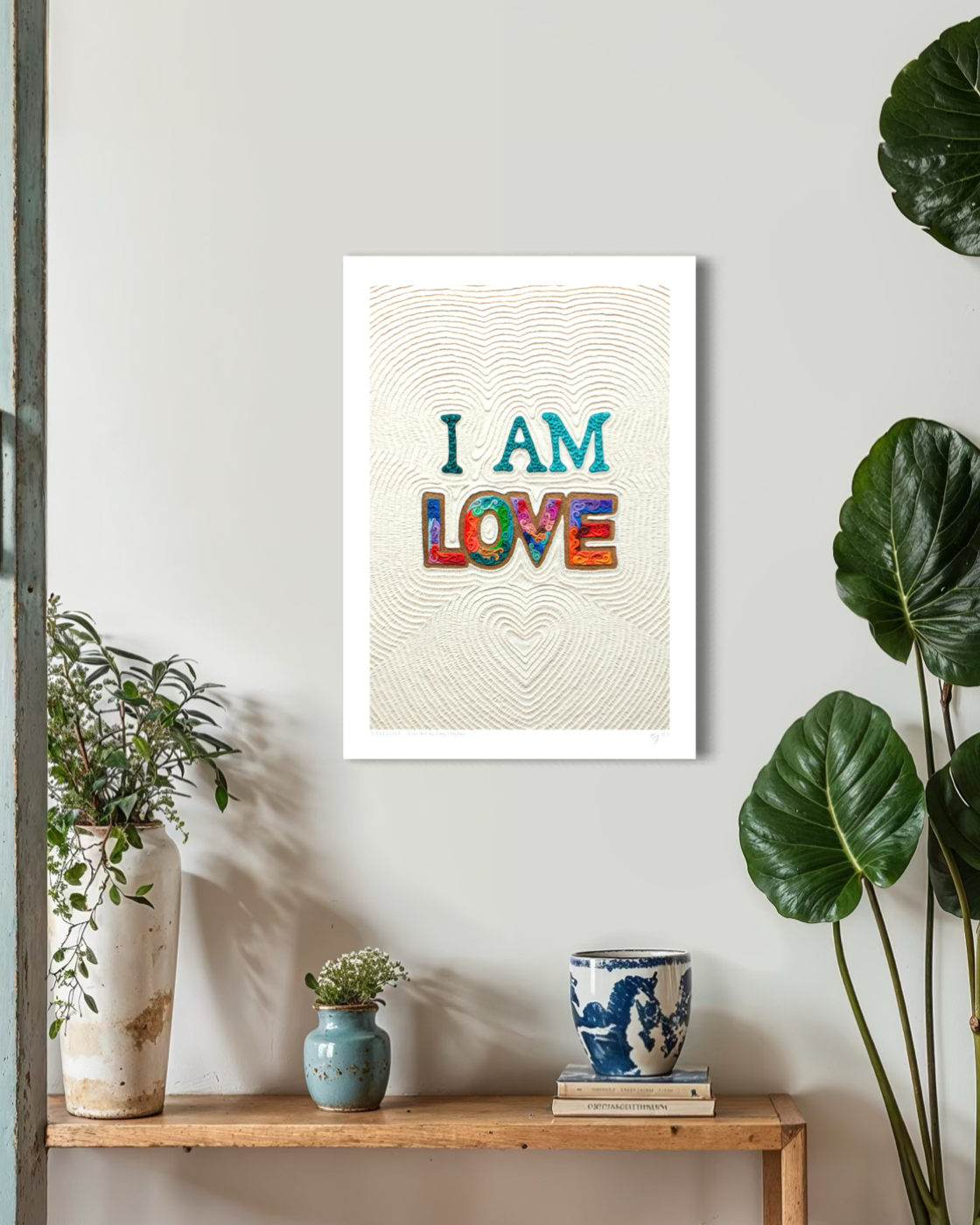An A3 wall fine art print with the words “I AM LOVE” in colorful textured letters hangs on a light gray wall above a wooden shelf with plants and small decor items. Art by New Zealand artist Emy Oikawa from Eclo Art.