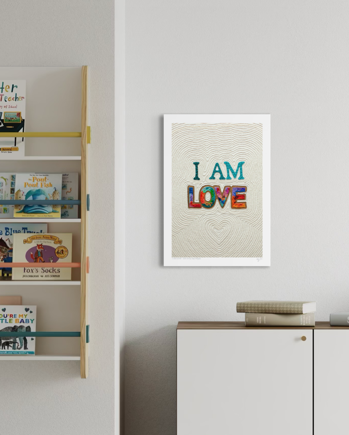 An A3 wall art print with the text 'I AM LOVE' in colorful letters on a textured cream background made originally with plasticine clay, hanging on a light gray wall in a styled room with a bookshelf and cabinet. Art by New Zealand artist Emy Oikawa from Eclo Art.