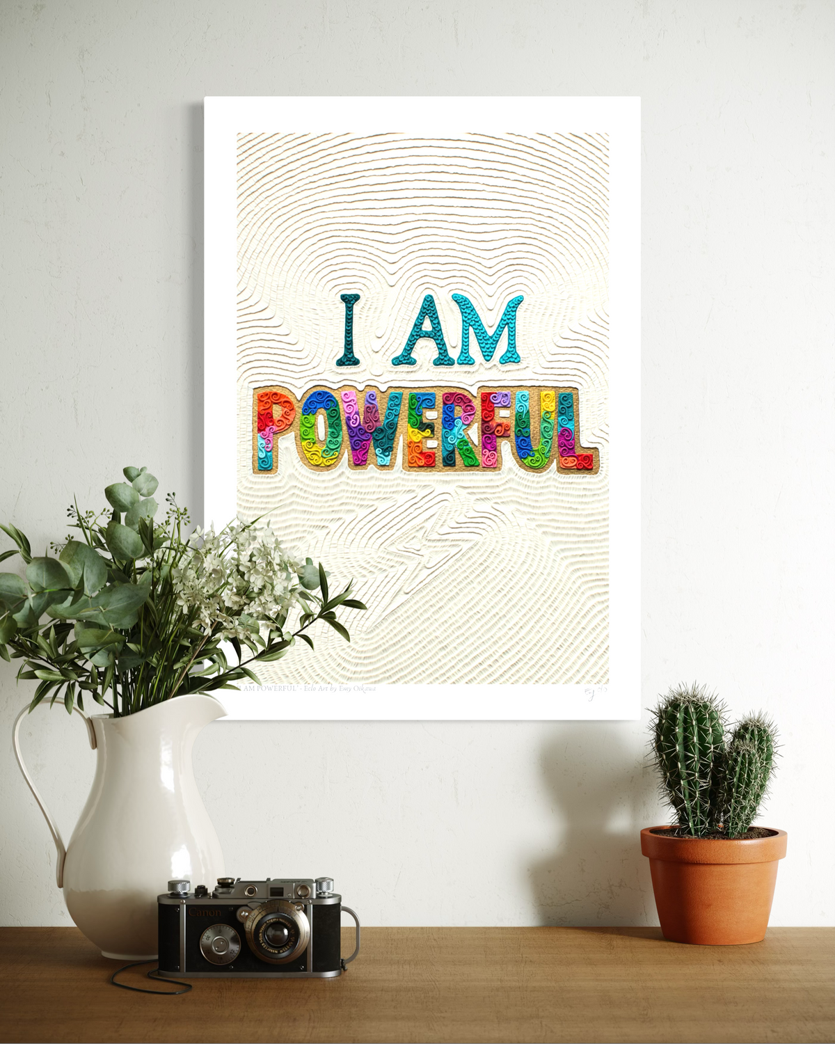 An A3  typographic wall art print reading 'I AM POWERFUL' Originally made using plasticine clay, hangs on a light wall above a wooden surface with a white pitcher of greenery, a vintage camera, and a small potted cactus. Art by New Zealand clay artist Emy Oikawa from Eclo Art.