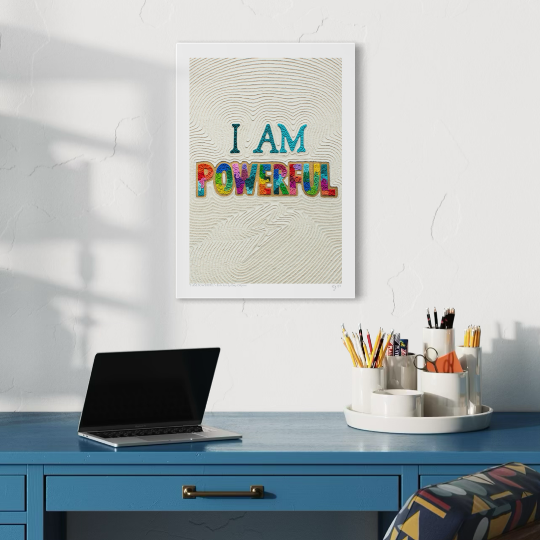 An A3 motivational fine art print with the text 'I AM POWERFUL' hangs on a white wall above a blue desk. The print features a textured beige background with a thunder light shape at the bottom made originally fully with plasticine clay. Art by New Zealand clay artist Emy Oikawa from Eclo Art.