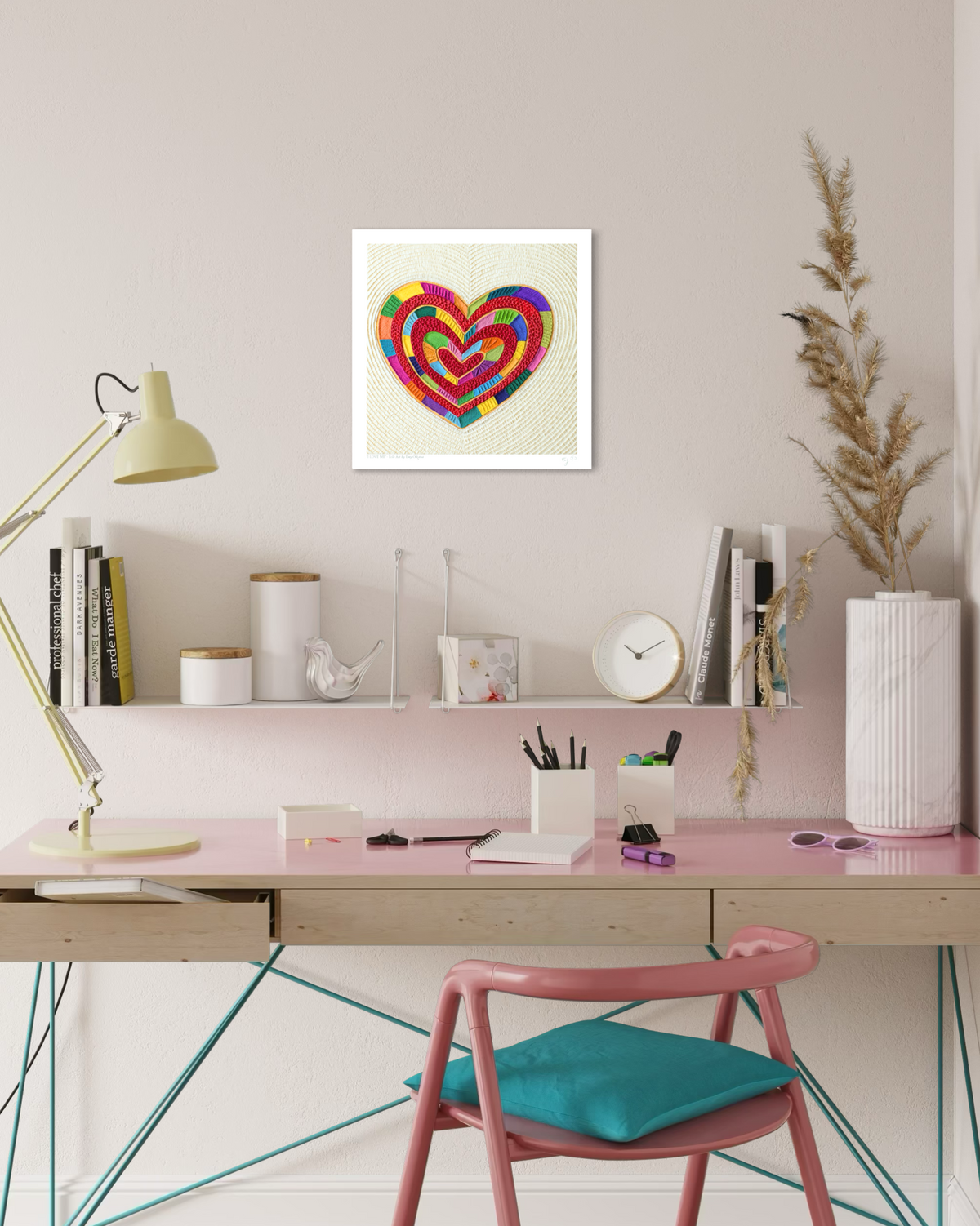 A colorful, multi-layered fine art print heart called ''I Love Me'' by clay artist Emy Oikawa, displayed on a light pink wall above a styled desk setup.