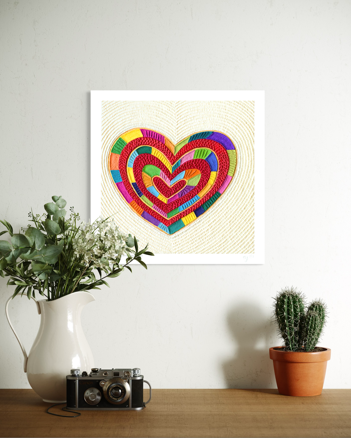 An 30 x 30 cm fine art print featuring a colorful, textured heart called ''I Love Me'' originally made of plasticine, displayed on a white wall above a wooden table some home decorations. By Clay Artist Emy Oikawa from Eclo Art.