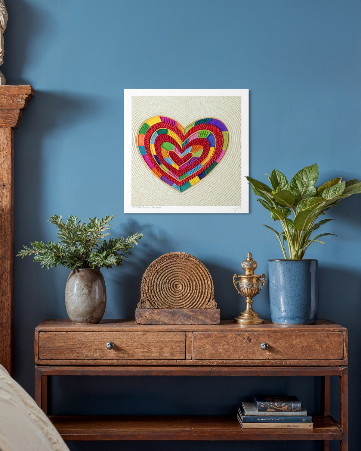 A fine art print featuring a colorful plasticine heart piece called ''I Love Me'' by Emy Oikawa, design on a beige textured background, hanging on a blue wall above a wooden console table with plants and decorative objects. New Zealand Artist from Eclo Art.