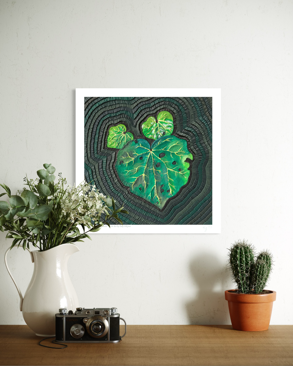 An art print called Kawakawa Leap of Faith, featuring a green leaf pattern on a dark textured background, originally made using plasticine clay, displayed on a white wall above a wooden table with a white pitcher of flowers, a vintage camera, and a small potted cactus. Art by artist Emy Oikawa from Eclo Art