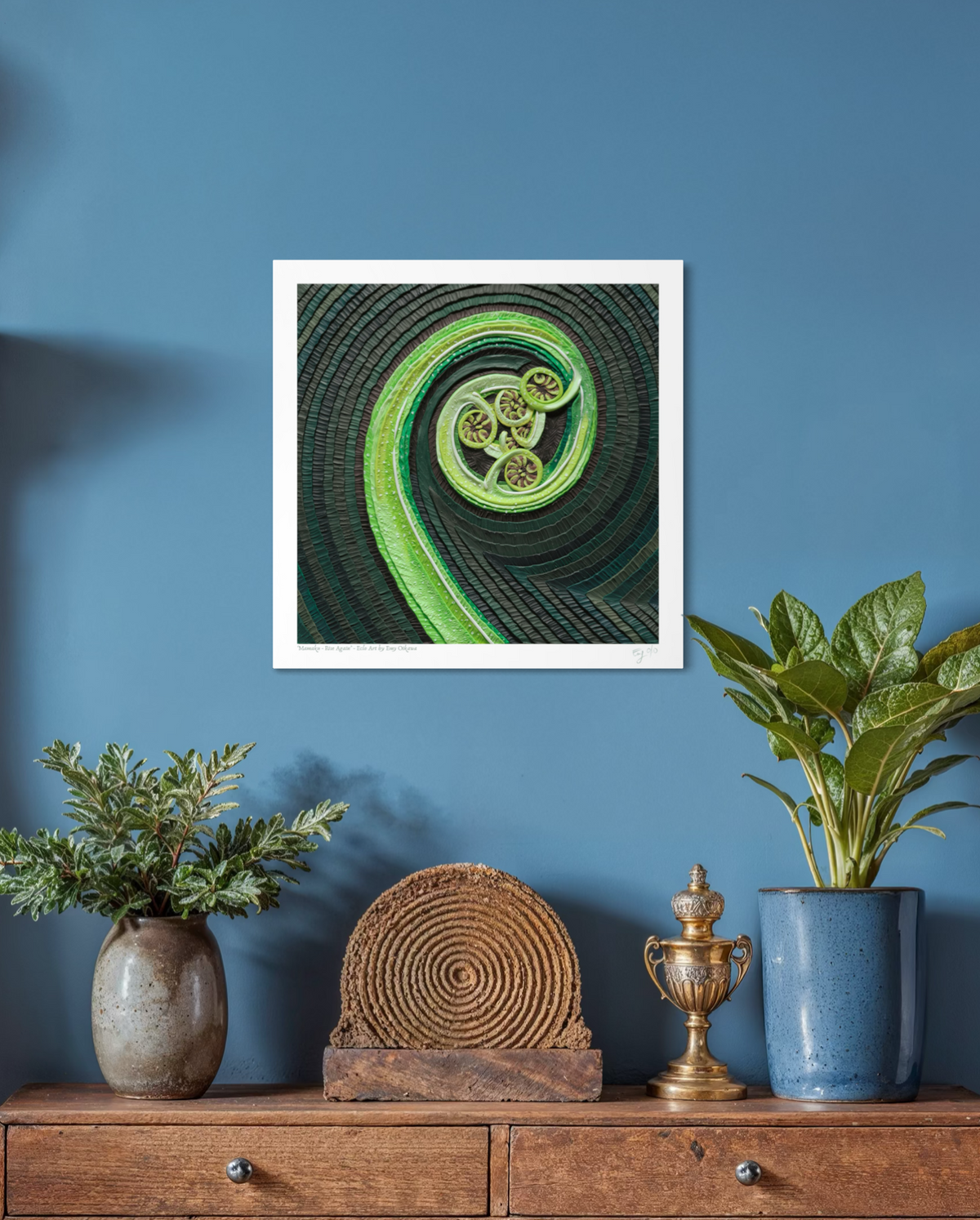 An art print of a New Zealand fern called Mamaku, featuring a green spiral design on a dark textured background, originally made using plasticine clay displayed on a blue wall above a wooden console table with plants and decorative objects.  Art by clay artist Emy Oikawa from Eclo Art. New Zealand nature.