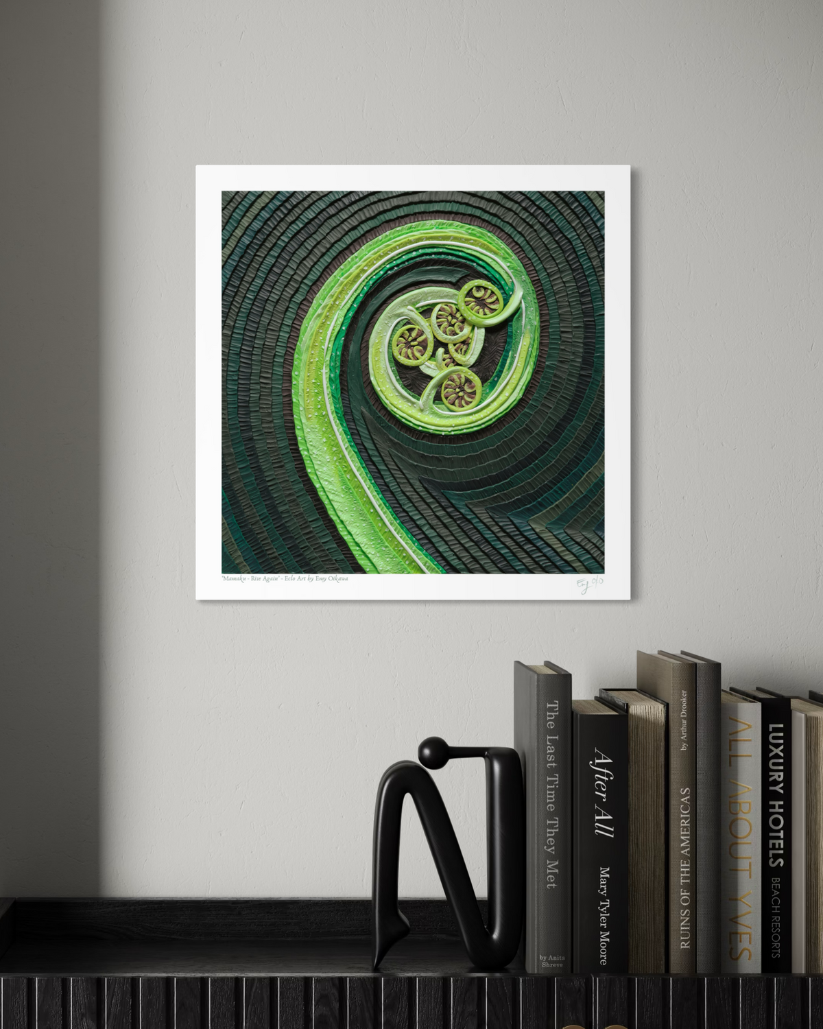An art print featuring The Mamaku New Zealand Fern, a green spiral plant, originally made with plasticine clay, displayed on a light gray wall above a shelf with books and a black decorative object. Art by clay artist Emy Oikawa from Eclo Art. New Zealand nature.
