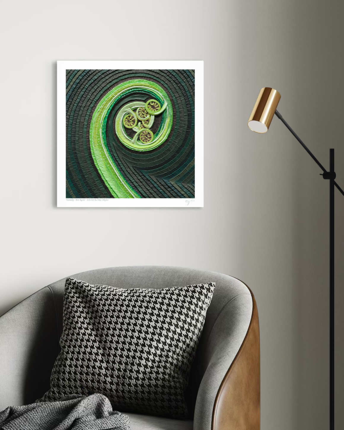 An art print of a New Zealand fern called Mamaku, featuring a green and black spiral design, displayed on a light gray wall above a gray armchair with a black-and-white houndstooth pillow and a gold floor lamp nearby.  Art by clay artist Emy Oikawa from Eclo Art. New Zealand nature.