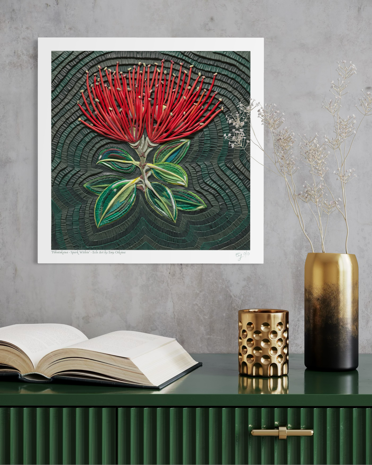 A botanical art print of a red New Zealand flower called Pohutukawa, with green leaves on a dark textured background, all made originally with plasticine, displayed on a dark green cabinet against a gray wall with decorative props including an open book, a gold vase, and dried stems. Art print by New Zealand artist Emy Oikawa, Eclo Art.