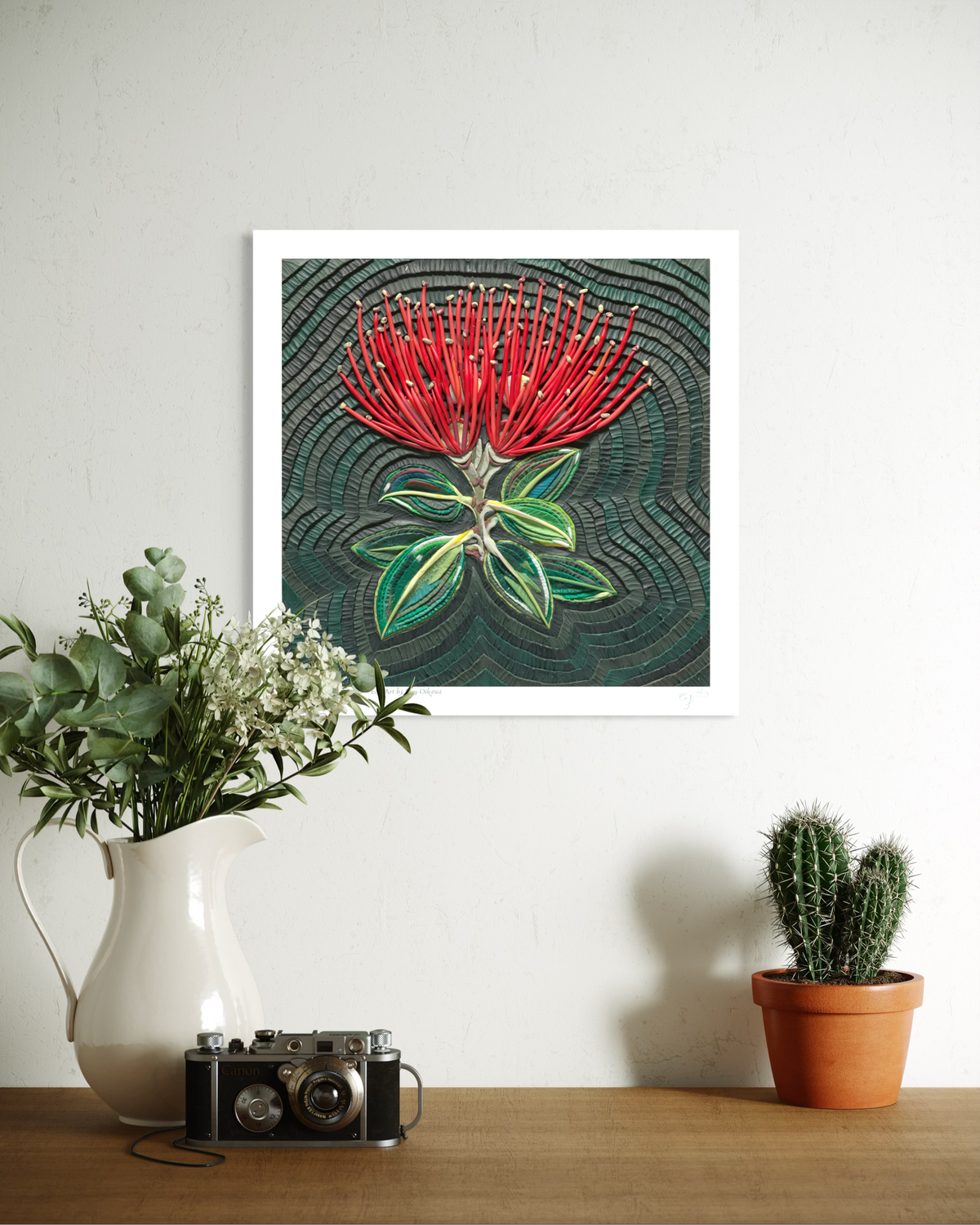 A wall art print featuring a red flower from New Zealand called Pohutukawa, with green leaves on a dark textured background, displayed on a white wall above a wooden table with a white pitcher of flowers, a vintage camera, and a small potted cactus. Pohutukawa Spark Within plasticine clay art print by New Zealand artist Emy Oikawa from Eclo Art.