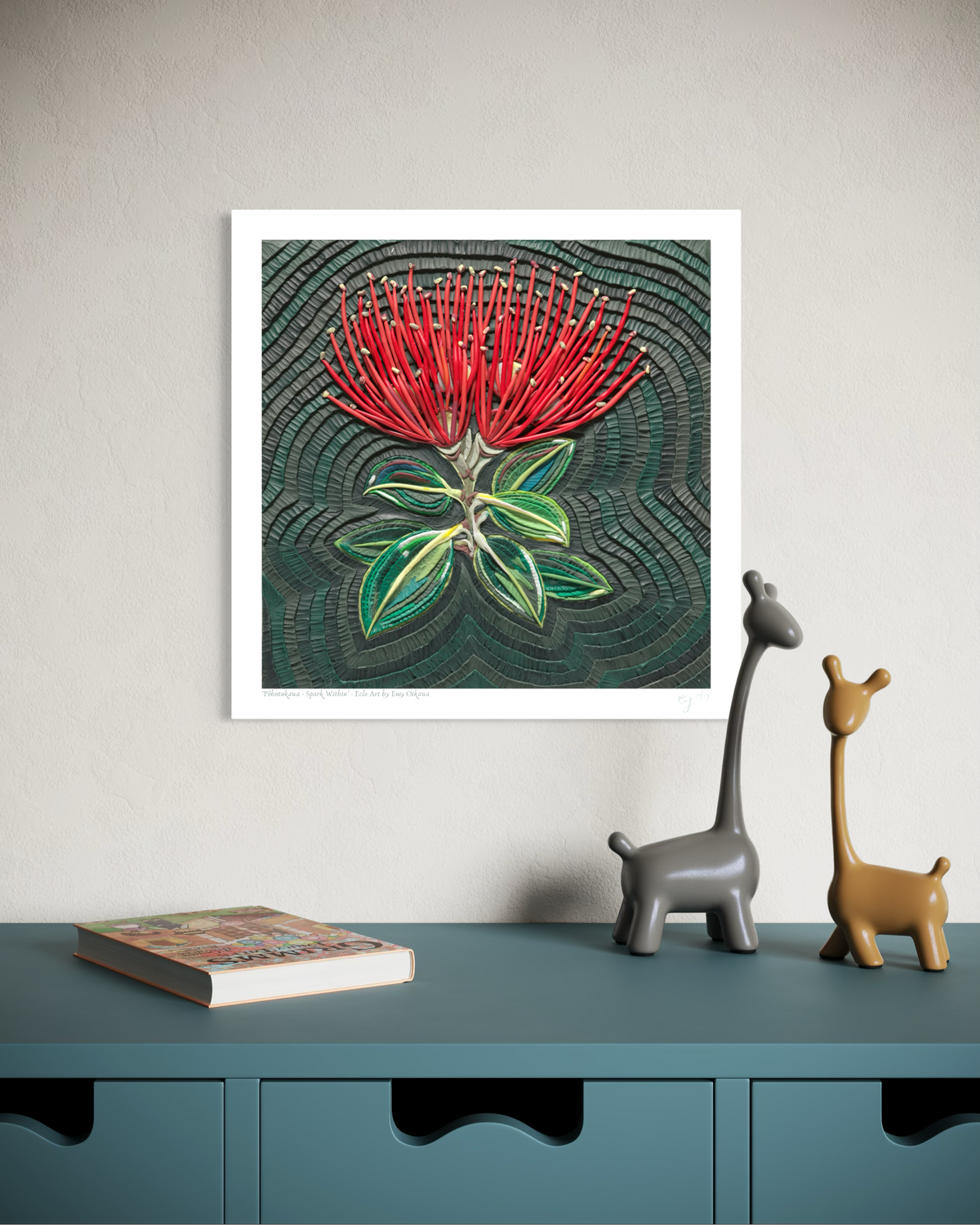 A wall art print featuring a red flower called Pohutukawa with green leaves on a dark textured background, originally made with Plasticine clay, displayed above a teal cabinet with two small giraffe figurines and a book. Pohutukawa Spark Within plasticine clay art print by New Zealand artist Emy Oikawa from Eclo Art.