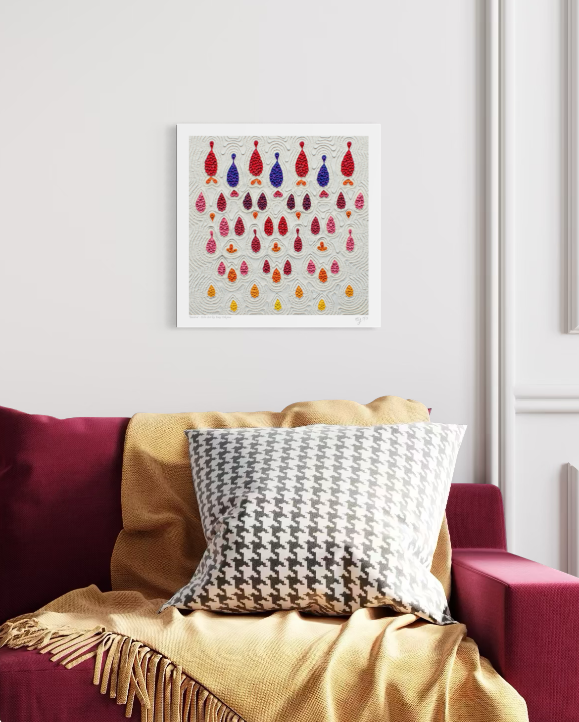 An abstract art print called ''Receive'' featuring colorful hanging teardrop-shaped plasticine on a light gray plasticine background, displayed on a white wall above a red sofa with a yellow throw blanket and a black-and-white patterned pillow. Art by New Zealand clay artist Emy Oikawa.