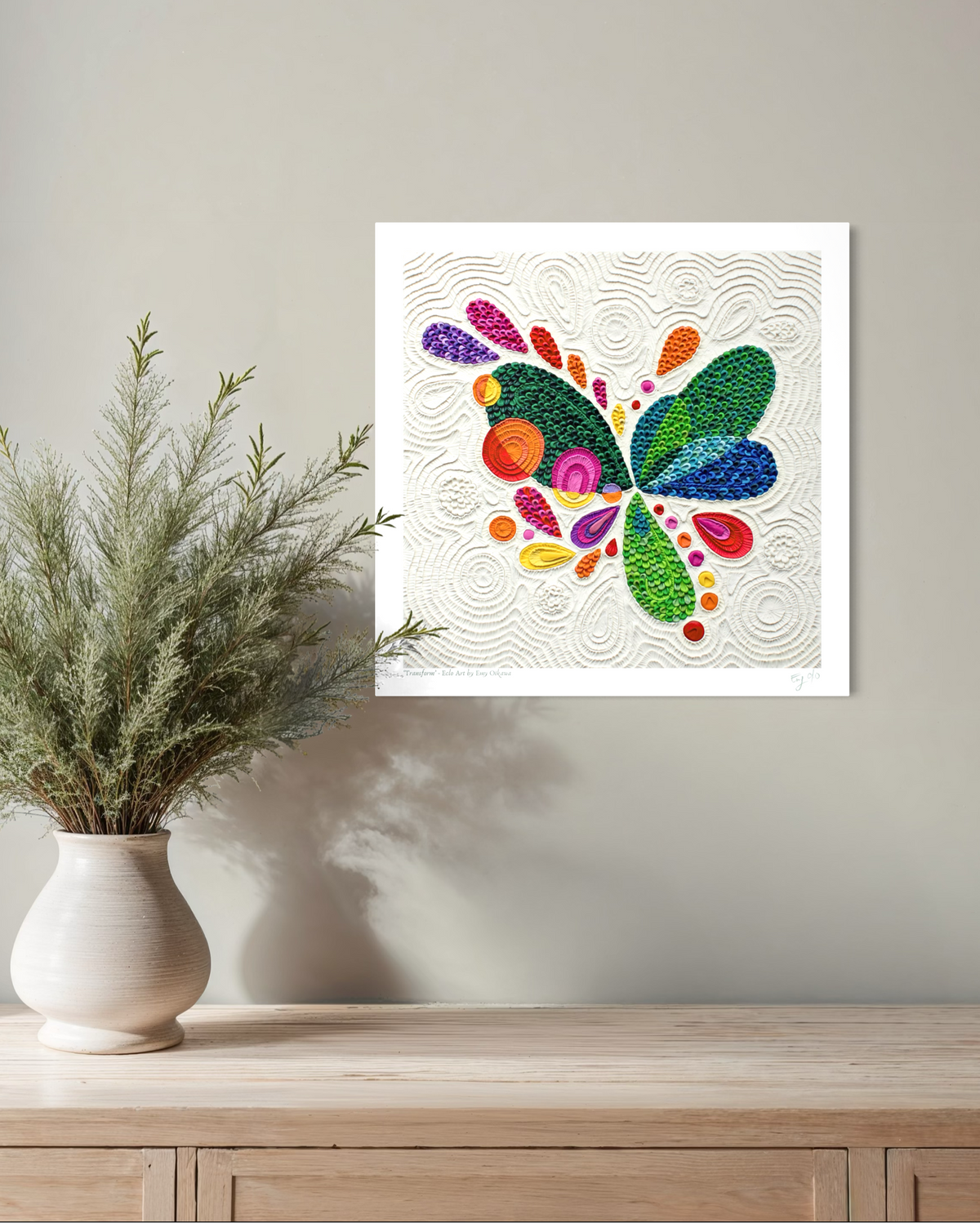A square wall art print called ''Transform'' featuring a colorful abstract butterfly design on a textured off-white plasticine background, displayed on a light gray wall above a wooden console table with a white vase of greenery.