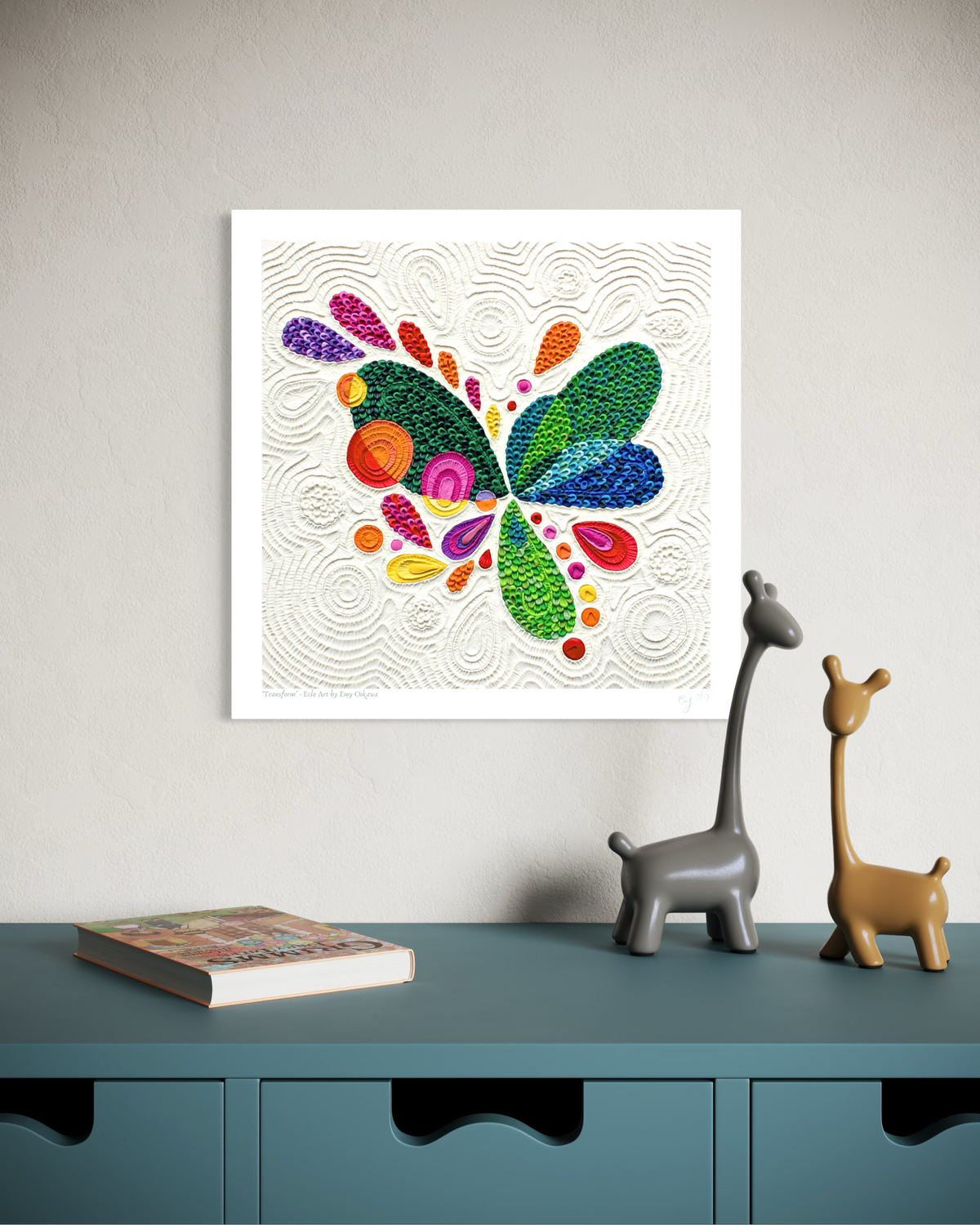 A wall fine art print called ''Transform'' featuring a colorful abstract butterfly made of textured plasticine on a white background, displayed above a teal cabinet with two small giraffe figurines and a book. Art by clay artist Emy Oikawa from Eclo Art.
