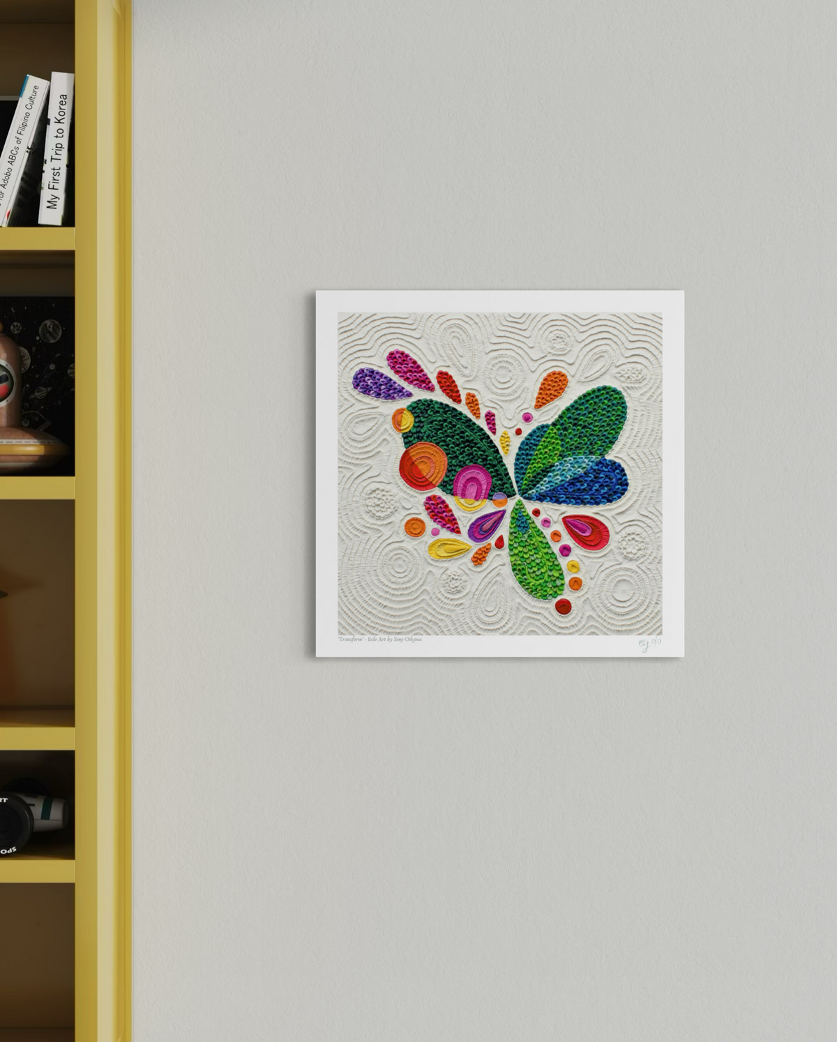 A square wall art print featuring a colorful butterfly abstract, originally made of plasticine drop shapes in green, blue, purple, pink, orange, yellow, and red, displayed on a light gray wall next to a yellow bookshelf. Art by clay artist Emy Oikawa from Eclo Art.