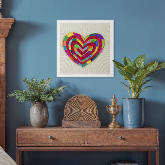 video close up of a fine art print featuring a colorful plasticine heart piece called ''I Love Me'' by Emy Oikawa, design on a beige textured background, hanging on a blue wall above a wooden console table with plants and decorative objects. New Zealand Artist from Eclo Art.