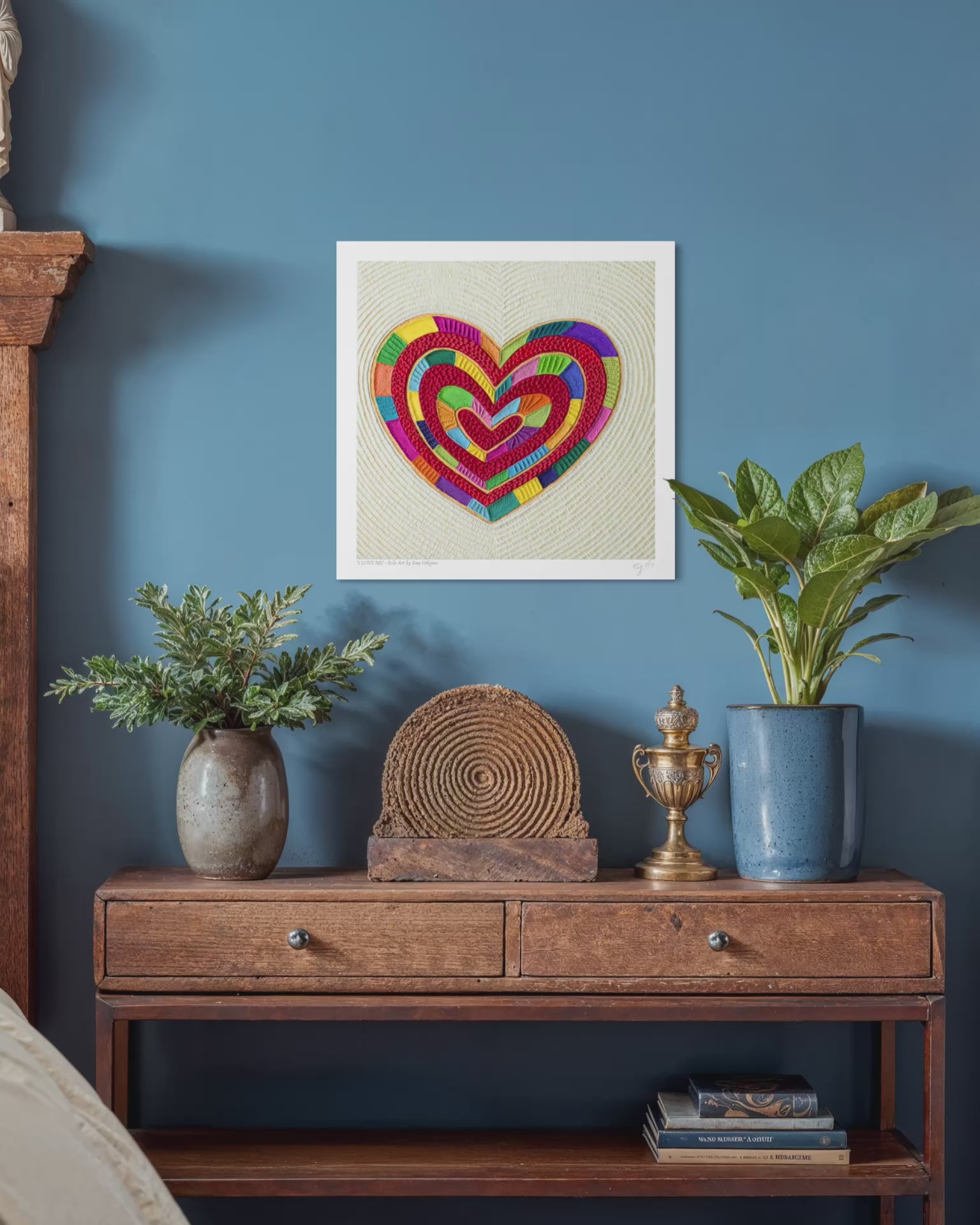 video close up of a fine art print featuring a colorful plasticine heart piece called ''I Love Me'' by Emy Oikawa, design on a beige textured background, hanging on a blue wall above a wooden console table with plants and decorative objects. New Zealand Artist from Eclo Art.