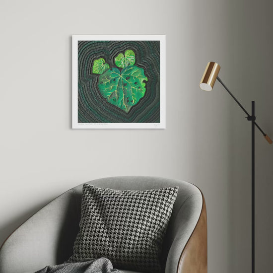 close up video of An art print featuring a green Kawakawa leaf pattern on a dark textured background, originally made using palsticine clay, displayed on a light gray wall above a gray armchair with a black-and-white patterned pillow and a floor lamp nearby. Art by artist Emy Oikawa from Eclo Art