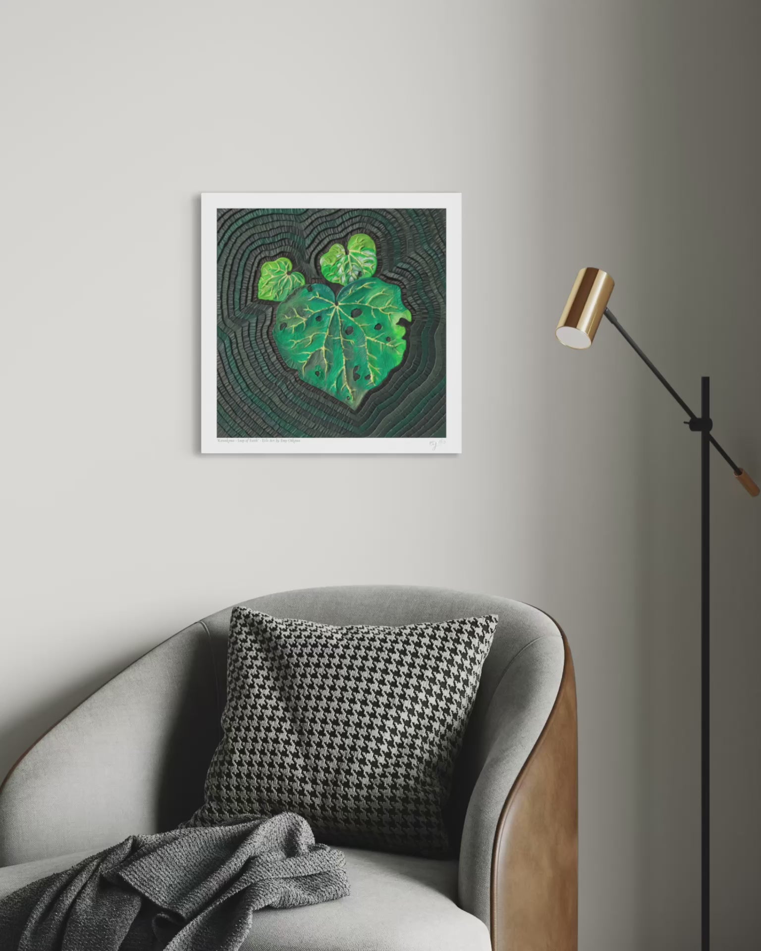 close up video of An art print featuring a green Kawakawa leaf pattern on a dark textured background, originally made using palsticine clay, displayed on a light gray wall above a gray armchair with a black-and-white patterned pillow and a floor lamp nearby. Art by artist Emy Oikawa from Eclo Art