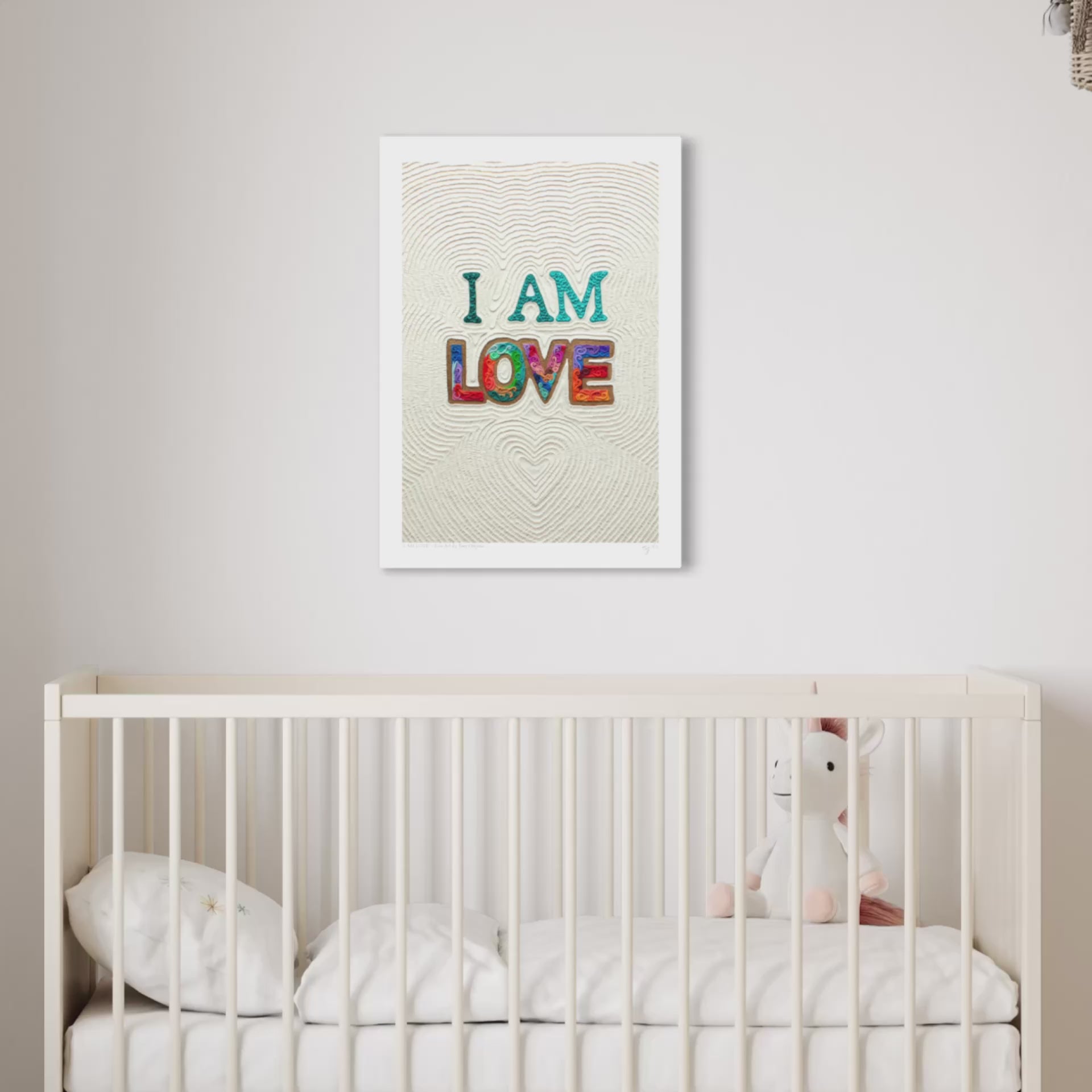 close up video of an A3 wall art print with the words 'I AM LOVE' in colorful textured letters hangs above a white crib in a softly lit nursery. Art by New Zealand artist Emy Oikawa from Eclo Art.
