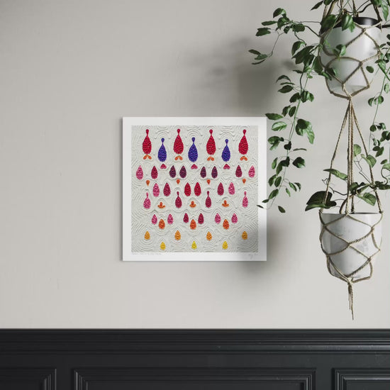 A square wall art print called ''Receive'' featuring rows of colorful hanging teardrop-shaped made with plasticine clay on a light gray background, displayed on a beige wall above a black wainscoted panel with a green hanging plant to the right. Art by New Zealand clay artist Emy Oikawa.