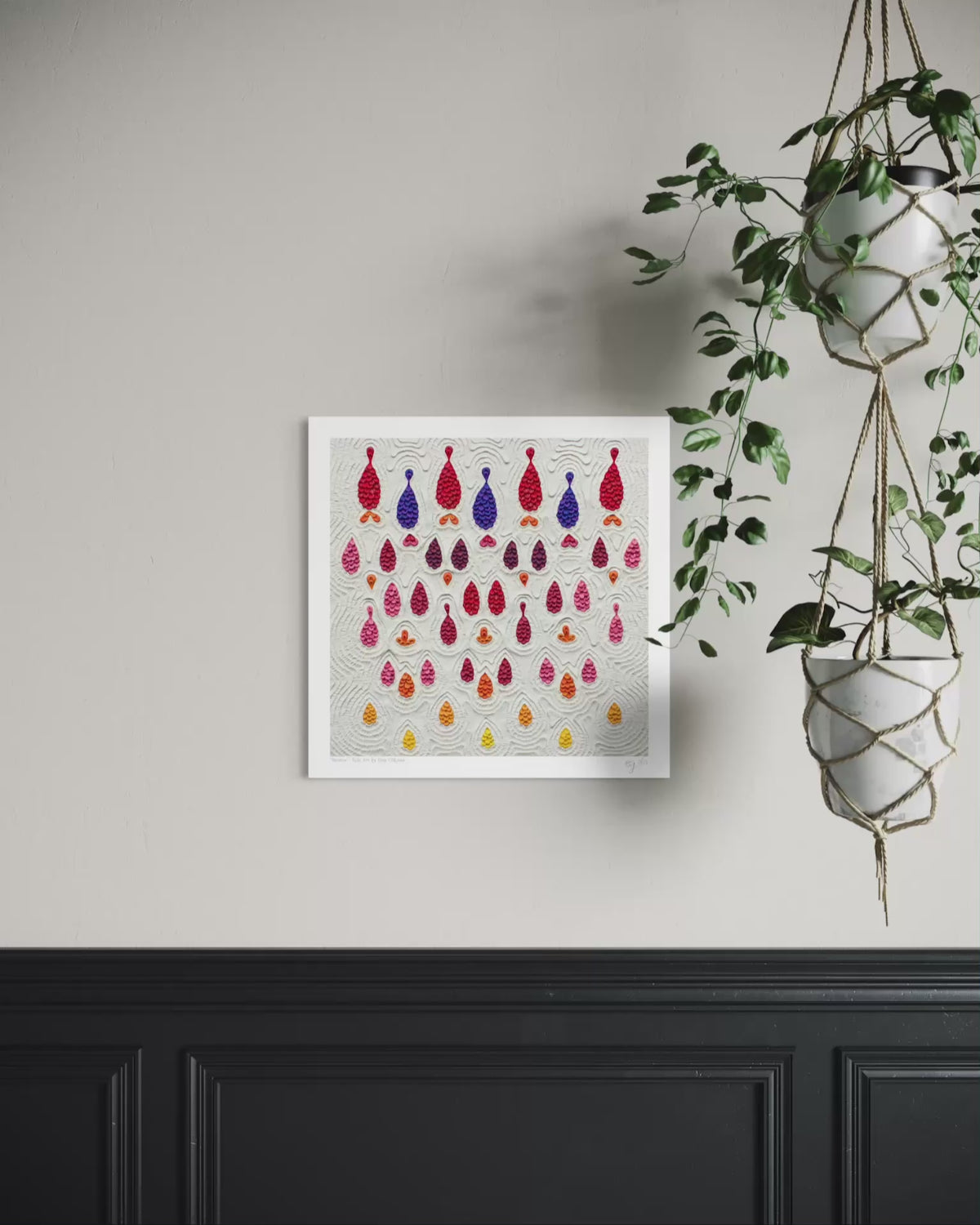 A square wall art print called ''Receive'' featuring rows of colorful hanging teardrop-shaped made with plasticine clay on a light gray background, displayed on a beige wall above a black wainscoted panel with a green hanging plant to the right. Art by New Zealand clay artist Emy Oikawa.