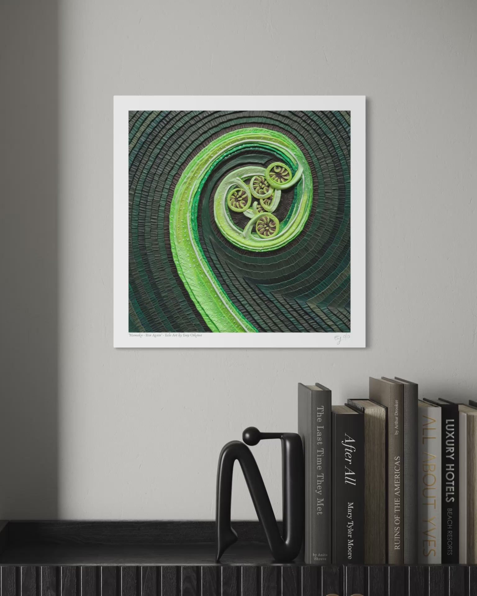 close up video of An art print featuring The Mamaku New Zealand Fern, a green spiral plant, originally made with plasticine clay, displayed on a light gray wall above a shelf with books and a black decorative object. Art by clay artist Emy Oikawa from Eclo Art. New Zealand nature.