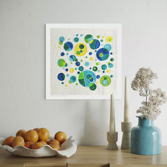close up video of A square fine wall art print called ''Abundance'' featuring colorful abstract circles in blue, green, yellow on a cream plasticine colour background, displayed above a wooden console table with oranges, candles, and a blue vase in a styled interior setting. By New Zealand clay artist Emy Oikawa from Eclo Art.