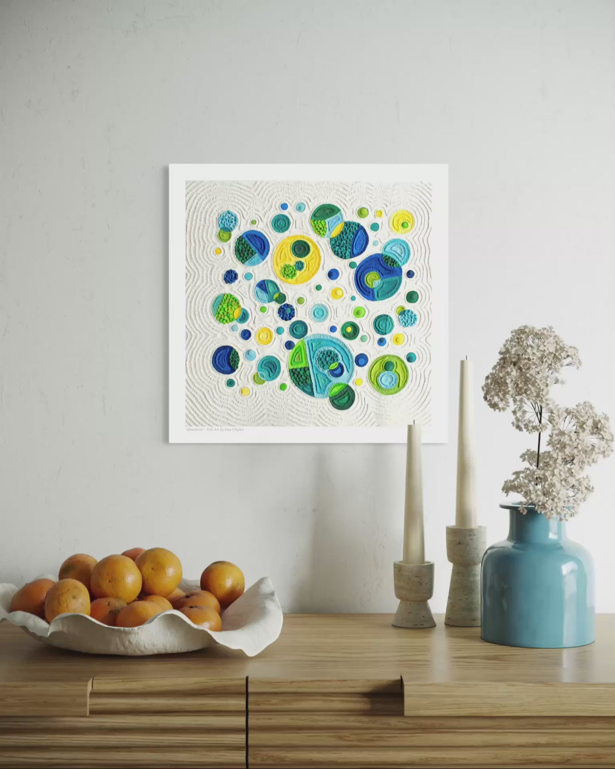 close up video of A square fine wall art print called ''Abundance'' featuring colorful abstract circles in blue, green, yellow on a cream plasticine colour background, displayed above a wooden console table with oranges, candles, and a blue vase in a styled interior setting. By New Zealand clay artist Emy Oikawa from Eclo Art.