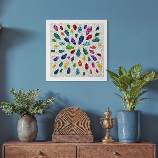 close up video of A square plasticine wall art print called ''Empower'' featuring a colorful radial pattern teardrop shapes on a white background, displayed on a blue wall above a wooden console table with plants and decorative objects. Art by New Zealand Artist Emy Oikawa from Eclo Art.