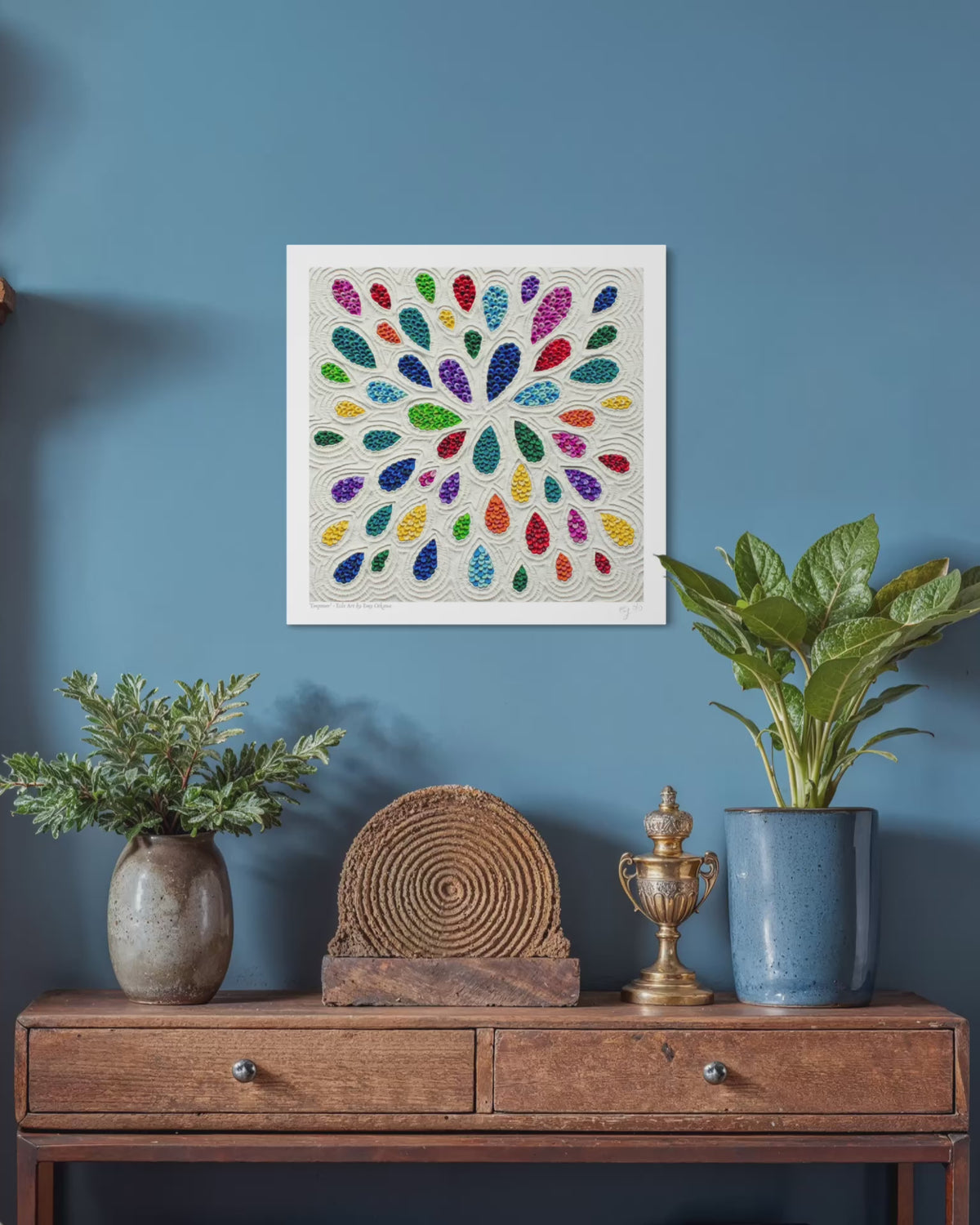 close up video of A square plasticine wall art print called ''Empower'' featuring a colorful radial pattern teardrop shapes on a white background, displayed on a blue wall above a wooden console table with plants and decorative objects. Art by New Zealand Artist Emy Oikawa from Eclo Art.