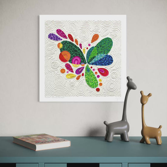 Video close up of a wall fine art print called ''Transform'' featuring a colorful abstract butterfly made of textured plasticine on a white background, displayed above a teal cabinet with two small giraffe figurines and a book. Art by New Zealand clay artist Emy Oikawa from Eclo Art.