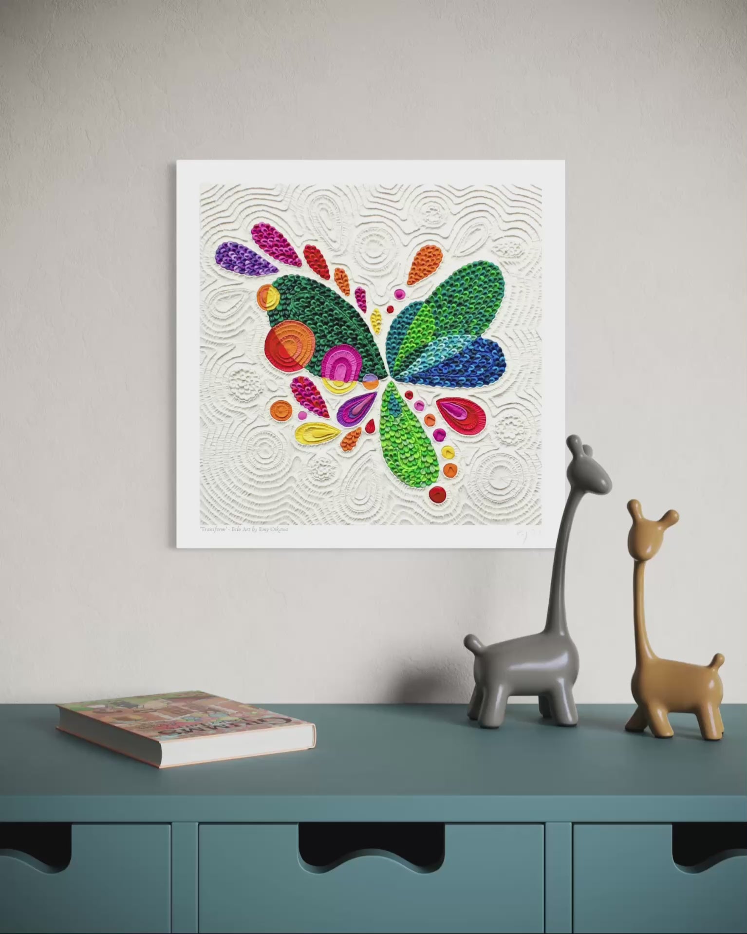 Video close up of a wall fine art print called ''Transform'' featuring a colorful abstract butterfly made of textured plasticine on a white background, displayed above a teal cabinet with two small giraffe figurines and a book. Art by New Zealand clay artist Emy Oikawa from Eclo Art.